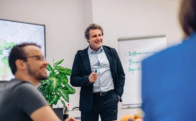 Gereon Friederes steht vor einem Flipchart und spricht zu einer Gruppe von Menschen in einem Besprechungsraum. Auf dem Flipchart stehen die Begriffe Differenzierung, Glaubwürdigkeit und Begeisterung.