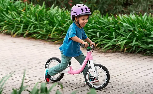 Kind fährt mit Helm auf einem pinken woom-Laufrad über einen gepflasterten Weg in grüner Umgebung. Bild steht für Kinderfahrräder, sichere Mobilität und zielgruppengerechte Markenkommunikation.